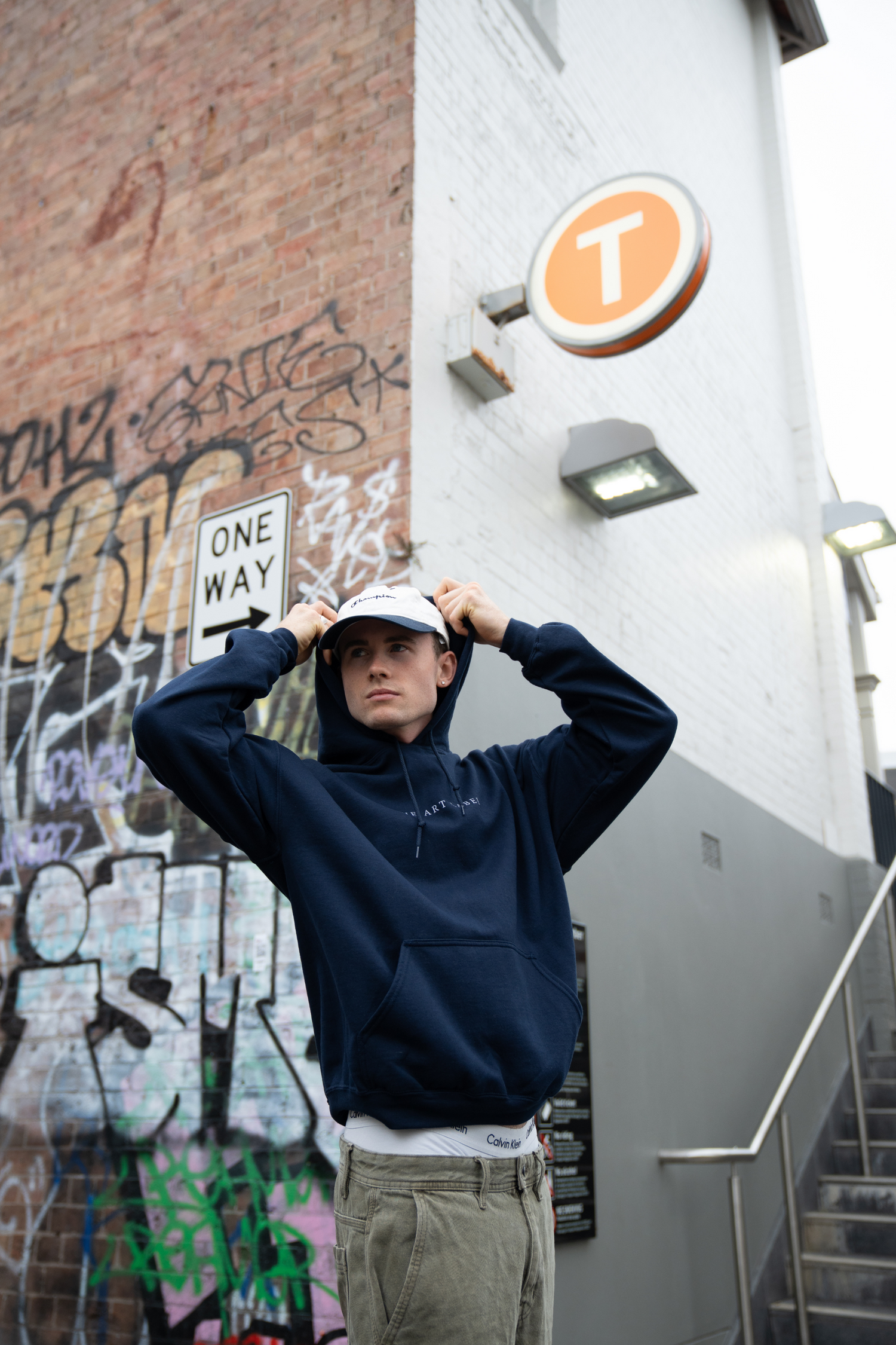 A guy standing on a corner close to a Sydney train station holding his hood up and looking around