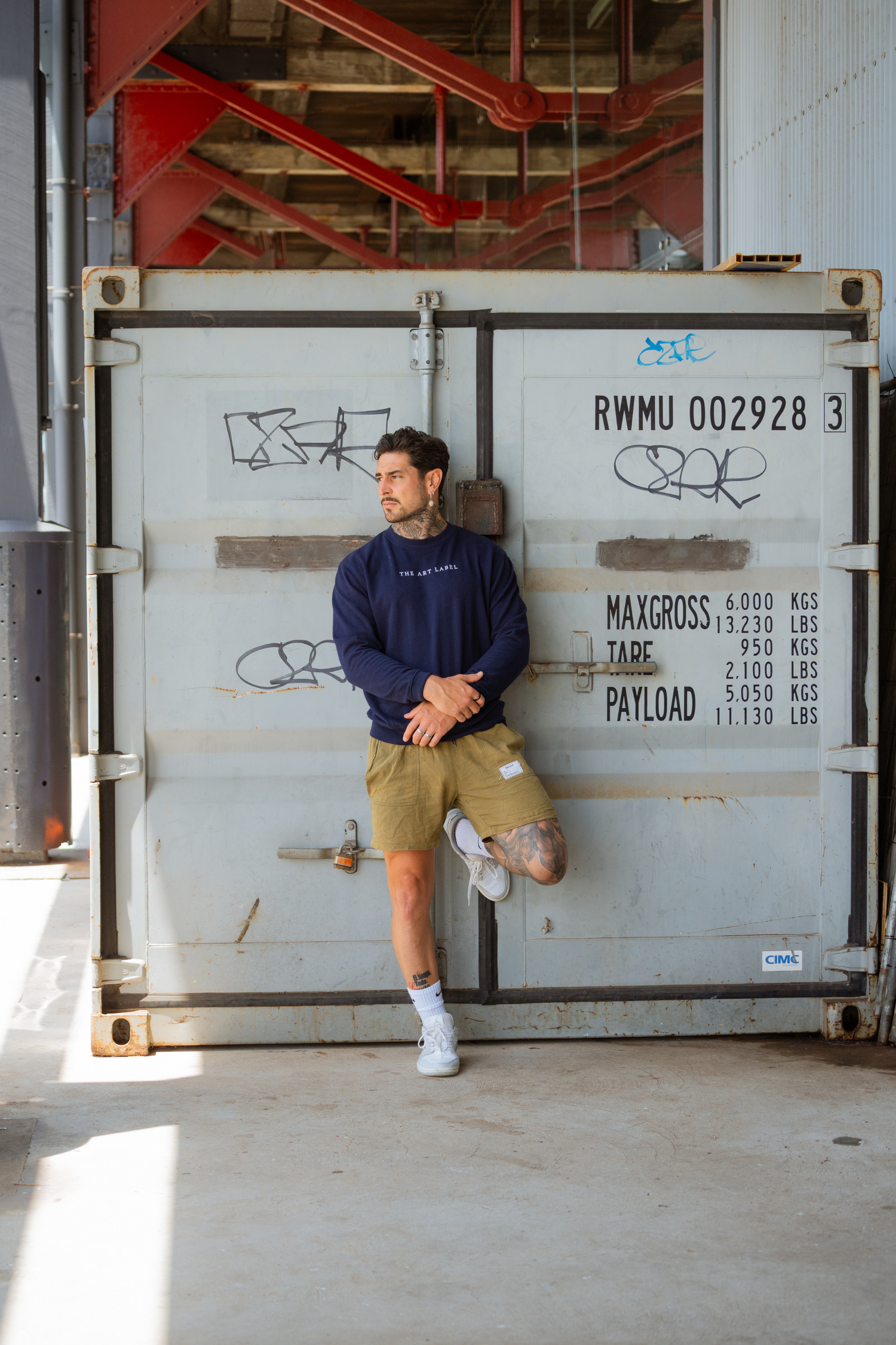 A guy leaning with the back to a container, one foot up against the container wall and looking to the side wearing wearable art